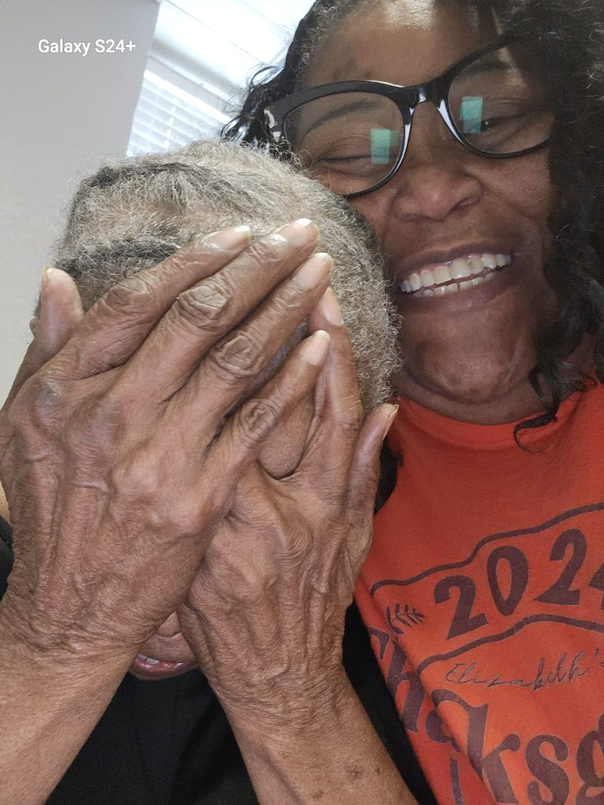 The woman covering her face with both hands as she laughs, pressed cheek to cheek with our founder, who is smiling broadly in glasses and an orange shirt