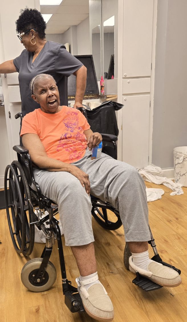 The woman in a wheelchair wearing an orange shirt and gray pants, laughing with her mouth wide open while a caregiver in gray scrubs stands behind her